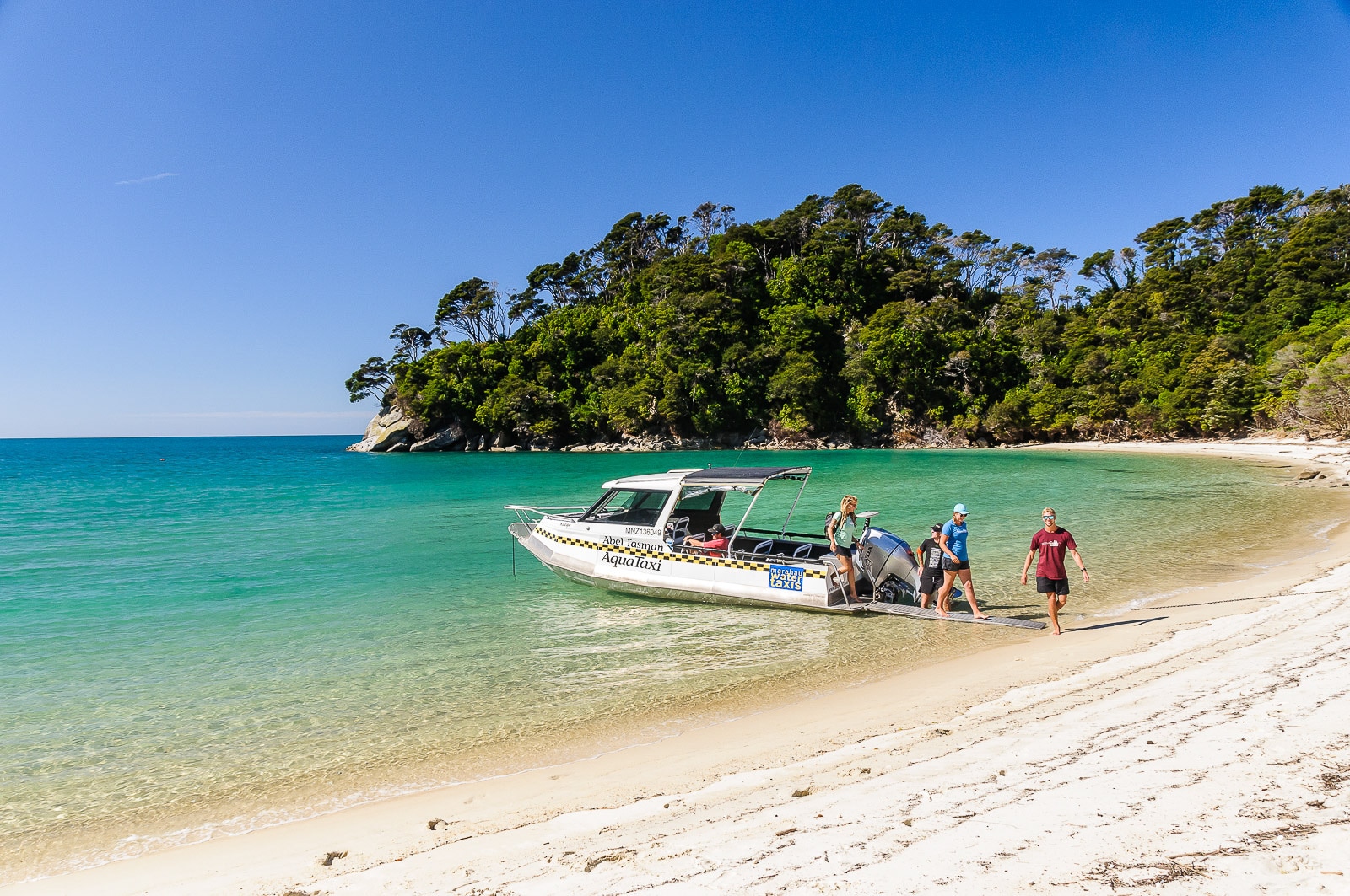 Aqua-taxi shuttle offloading travelers onto a golden beach in Abel Tasman National Park, New Zealand, with clear waters and forested coastline in the background